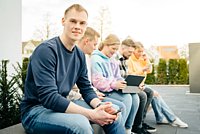 Werkstudenten sitzen auf einer Terrasse. Einer guckt in die Kamera, die restlichen auf ein mobiles Endgerät. 