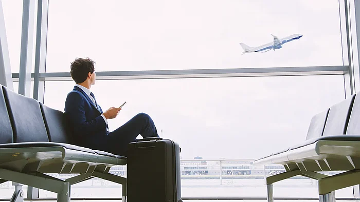 Mann im Anzug sitzt mit Koffer am Gate und blickt einem startenden Flugzeug nach
