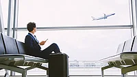 Mann im Anzug sitzt mit Koffer am Gate und blickt einem startenden Flugzeug nach
