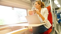 Frau sitzt im Zug an einem Platz mit einem Laptop vor sich und hat währenddessen ein Smartphone in der Hand. 