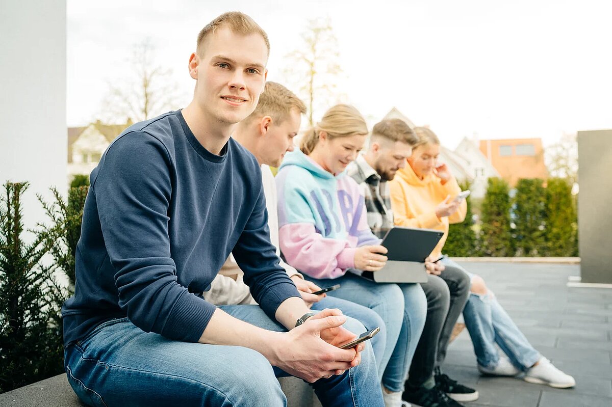 Werkstudenten sitzen auf einer Terrasse. Einer guckt in die Kamera, die restlichen auf ein mobiles Endgerät. 