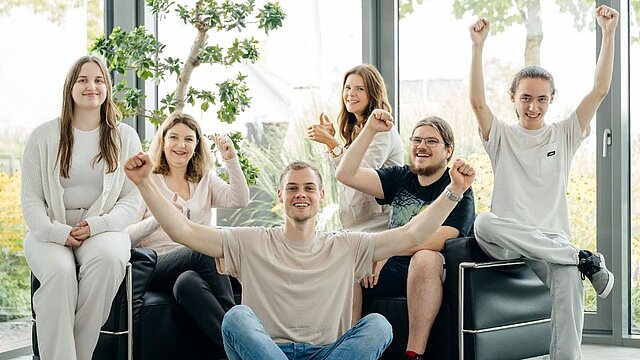 Ein junger Mann sitzt im Schneidersitz im Vordergrund, eine Gruppe befindet sich dahinter. Sie jubeln.