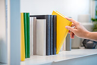 Bücher in einem Buchregal. Ein gelbes Buch ist leicht gekippt und wird aus dem Regal herausgenommen.