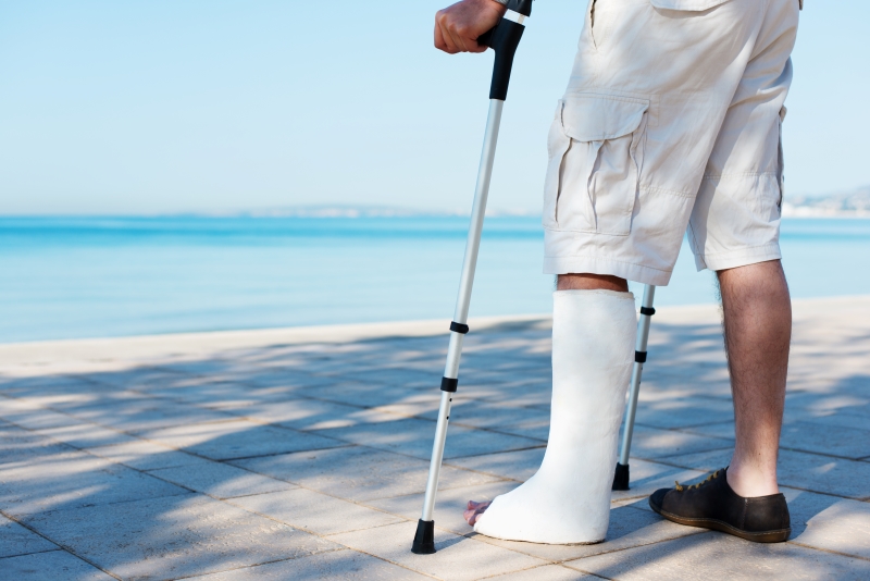 Person mit Krücke und Schiene am Bein steht am Strand